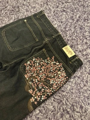 Svarta derschutze jeans med broderat körsbärsträd - Unika svarta jeans från derschutE med ett snyggt broderat körsbärsträd i rosa och vitt på bakfickan. Jeansen har raka ben och klassisk femficksmodell. Perfekta för dig som vill sticka ut med coola detaljer och kreativ design.