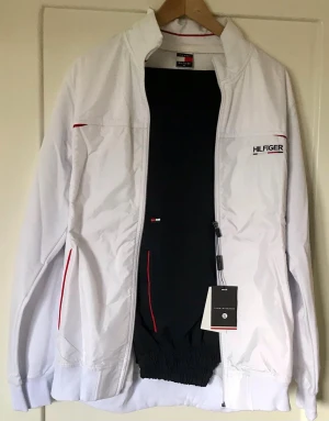 Vit vindjacka med byxor  från Tommy Hilfiger - Snygg vit vindjacka med byxor  från Tommy Hilfiger med dragkedja, ståkrage och diskreta röda detaljer. Jackan har logotypen broderad på bröstet och elastiska muddar. Perfekt för dig som vill ha en stilren och sportig look.