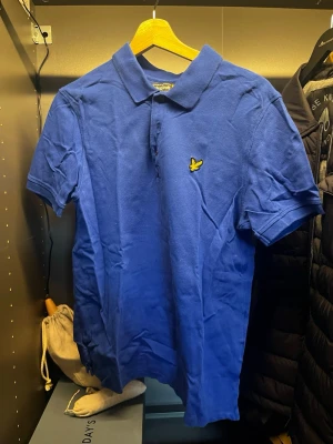 Blå pikétröja från Lyle & Scott - Säljer en klassisk blå pikétröja från Lyle & Scott med korta ärmar och den ikoniska gula logon på bröstet. Tröjan har krage och knappar framtill, perfekt för en clean och stilren look.