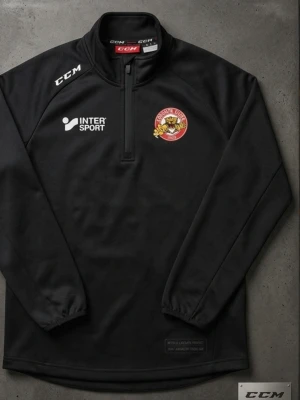 Svart träningströja från CCM - Svart långärmad träningströja från CCM med halv dragkedja, klubbmärke från Rögle BK på bröstet och vita loggor från Intersport och CCM. Tröjan har resår vid ärmsluten och initialerna FB tryckta nedtill. Perfekt för hockeyträning eller uppvärmning.