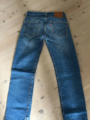 Levi's blå raka jeans - Klassiska blå Levi's jeans med rak passform och fem fickor. Jeansen har en vintage tvätt och tydliga sömmar, samt den ikoniska röda Levi's-lappen på bakfickan. Tillverkade i slitstark denim, perfekta för en avslappnad och tidlös stil.