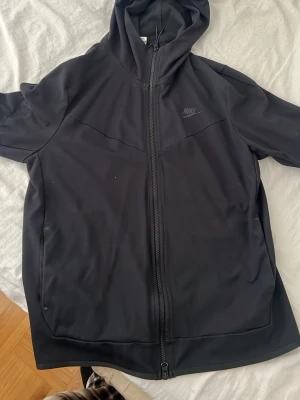 Svart zip hoodie från Nike - Säljer en svart hoodie från Nike med dragkedja framtill och diskret Nike-logga på bröstet. Hoodien har huva och är tillverkad i ett mjukt, stretchigt material som känns sportigt och bekvämt. Perfekt för dig som gillar en clean och stilren look.