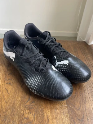 Svarta Puma fotbollsskor  - Säljer ett par svarta fotbollsskor från Puma med vita detaljer och klassisk logga på sidan. Skorna har snörning och slitstark ovandel i syntetmaterial samt fasta dobbar för bra grepp på planen. Perfekta för dig som vill ha stilrena och funktionella skor till matchen.