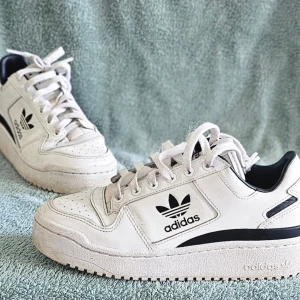 Vita Adidas sneakers med svart logga - Säljer ett par vita Adidas sneakers med svart Trefoil-logga på sidan och tungan. Skorna har klassisk låg profil, perforerad tå och svarta detaljer vid hälen. Tillverkade i läder med gummisula och vita skosnören. Perfekta för en clean och sportig stil.