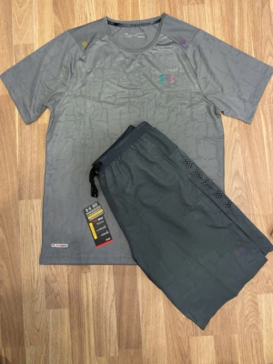 Grått träningsset från Under Armour - Snyggt grått träningsset från Under Armour med t-shirt och shorts. Setet har diskret mönster, ventilerande meshdetaljer på shortsen och logga på bröstet. Perfekt för gymmet eller löpning. Tillverkat i lätt och snabbtorkande material.
