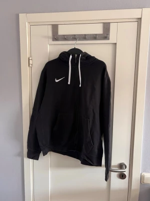 Svart Nike hoodie med dragkedja - Säljer en svart hoodie från Nike med vit logga på bröstet och vita snören i huvan. Hoodien har dragkedja framtill, två stora fickor och ribbade muddar. Perfekt för chill dagar eller när du vill ha något bekvämt och stilrent.