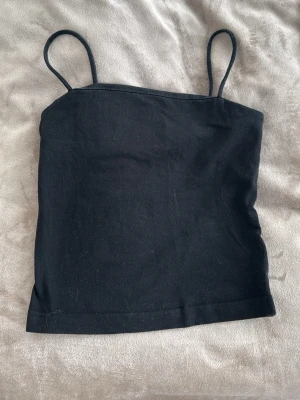 Svart enkel linnetopp - Säljer en svart, ärmlös linnetopp med smala axelband. Toppen har en rak och enkel design som passar perfekt till jeans eller kjol. Materialet känns mjukt och stretchigt, vilket gör den bekväm att bära hela dagen.