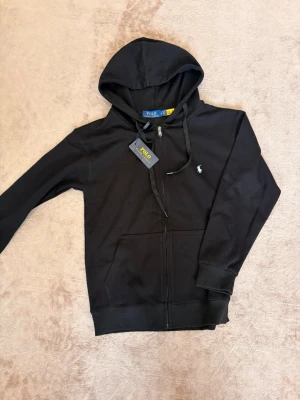 Svart hoodie från Polo Ralph Lauren - Svart hoodie med dragkedja från Polo Ralph Lauren. Klassisk design med huva, dragsko och två fickor framtill. Liten vit logga broderad på bröstet. Tillverkad i mjukt material som känns skönt mot huden. Perfekt för en avslappnad och stilren look.