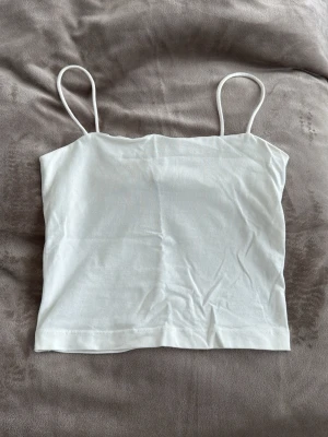 Vit enkel linnetopp med smala band - Säljer en vit linnetopp med tunna axelband och rak passform. Toppen är ärmlös och har en minimalistisk look som passar perfekt till sommaren. Tillverkad i mjukt bomullsmaterial för en bekväm känsla.