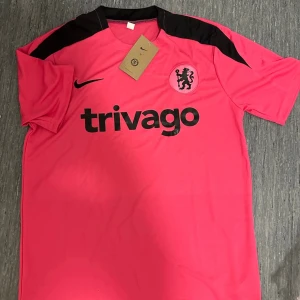 Rosa Chelsea tränings t-shirt Nike och shorts - Snygg rosa tränings t-shirt och shorts från Nike med Chelsea FC-logga och svart trivago-tryck på bröstet. Svarta detaljer på axlarna och klassisk rund hals. Perfekt för fotbollsträning eller gymmet.