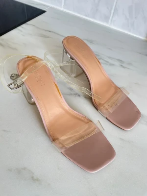 Beige sandaler med transparent klack - Säljer ett par trendiga sandaler från Truffle Collection med beige sula, fyrkantig tå och transparent rem över foten. Klacken är genomskinlig och ger en cool look. Remmarna är också transparenta och justerbara. Perfekta för att lyfta din outfit.