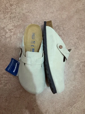 Birkenstock boston - Säljer ett par snygga beige tofflor från Birkenstock i mocka med korksula och svart räfflad yttersula. Klassisk slip-in modell med rem och knappdetalj på sidan. Perfekta för dig som gillar stilren och bekväm design.