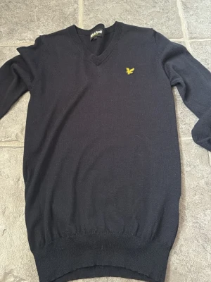 Mörkblå stickad tröja Lyle & Scott - Snygg mörkblå stickad tröja från Lyle & Scott med V-ringning och den klassiska gula logon broderad på bröstet. Tröjan har långa ärmar och ribbade muddar vid ärmslut och nederkant. Perfekt till lager på lager och enkel att matcha.