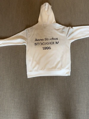 Vit hoodie från Acne Studios storlek S passar M - Snygg vit hoodie från Acne Studios med tryck på ryggen där det står 'Acne Studios STOCKHOLM 1996'. Klassisk modell med huva och ribbade muddar. Perfekt för en clean och stilren look. Materialet känns mjukt och skönt mot huden.