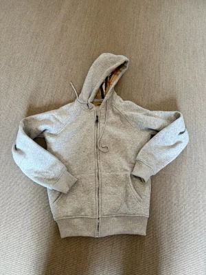 Grå hoodie från Burberry med dragkedja - Snygg grå hoodie från Burberry med klassiskt rutigt foder i huvan. Tröjan har dragkedja framtill, två fickor och ribbade muddar vid ärmslut och nederkant. Perfekt för en avslappnad och stilren look.