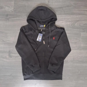 Svart hoodie från Polo Ralph Lauren - Svart hoodie med dragkedja från Polo Ralph Lauren. Klassisk design med huva och dragsnören, samt den ikoniska röda loggan broderad på bröstet. Tillverkad i mjukt bomullsmaterial och har ribbade muddar vid ärmslut och nederkant. Perfekt för en chill och stilren look. Tagg och påse ingår. Storlek M men skulle säga passar S bättre