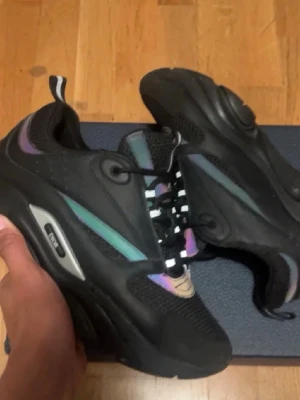 Svarta B22 sneakers med färgdetaljer - Säljer ett par svarta B2 sneakers med coola iriserande detaljer i grönt, lila och blått. Skorna har chunky sula, mesh och läderpaneler samt svartvita snören. Perfekta för dig som vill sticka ut med en unik och trendig look.