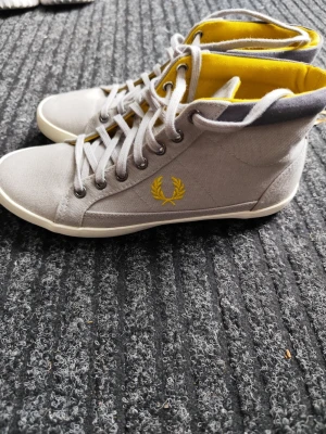 Gråa Fred Perry high top sneakers - Snygga high top sneakers från Fred Perry i grå canvas med gula detaljer på insidan och logga på sidan. Klassisk snörning och vit sula ger en clean look. Perfekta för dig som vill ha något stilrent men ändå färgglatt.