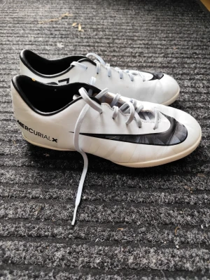 Nike Mercurial X CR7 fotbollsskor - Snygga vita Nike Mercurial X CR7 fotbollsskor med svart swoosh och detaljer. Skorna har lågt skaft, snörning och CR7-logga på hälen. Ovandelen är i syntetmaterial för bra bollkontroll och passform. Perfekta för dig som vill sticka ut på planen.