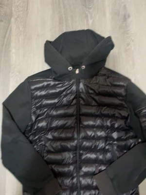 Svart dunjacka med huva från Moncler - Snygg svart dunjacka från Moncler med quiltad front och huva. Jackan har mjuka ärmar i sweatshirtmaterial och en ficka med Moncler-logga på ena ärmen. Dragkedja framtill och ribbade muddar. Perfekt för kalla vinterdagar.