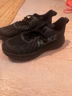 Svarta Hoka sneakers med mesh - Snygga svarta sneakers från Hoka med reflekterande logga på sidan. Skorna har en chunky sula och ovandel i meshmaterial för bra andningsförmåga. Klassisk snörning och sportig design som passar perfekt till streetwear.