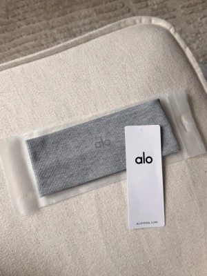 Grå ribbad hårband från Alo - Säljer ett stilrent grått hårband från Alo med diskret logga framtill. Hårbandet är ribbstickat och tillverkat i ett mjukt, stretchigt material som sitter bekvämt på huvudet. Perfekt för att hålla håret på plats under träning eller till vardags.