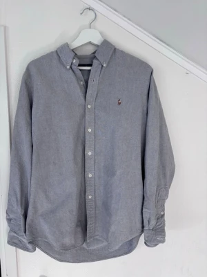 Grå skjorta från Ralph Lauren - Klassisk grå skjorta från Ralph Lauren i bomull med button-down krage och broderad logga på bröstet. Skjortan har långa ärmar, vita knappar och en tidlös, normal passform. Perfekt till både jeans och shorts för en clean look.                                Köptes inför sommaren och har använts mycket sparsamt, alltså fortfarande i väldigt bra skick. Kolla gärna in min profil för liknande plagg. Snabba affärer uppskattas 🙏