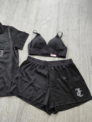Svart pyjamasset med strassdetaljer S  - Snyggt svart pyjamasset i satin med kortärmad skjorta, shorts och matchande bh-topp. Skjortan har knappar framtill och ficka med strassprydd bokstav. Shortsen har resår i midjan och samma strassdetalj. Perfekt för en lyxig hemmakväll. 