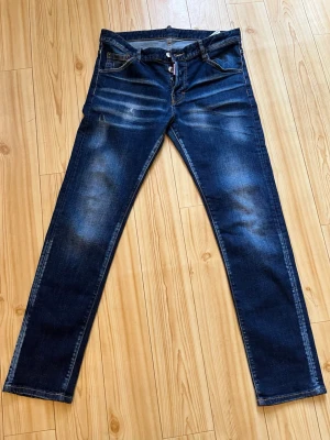 Dsquared2 - Säljer ett par mörkblå jeans med raka ben och snygga slitningar på låren och knäna. Jeanseno har klassisk femficksmodell och knäppning med knappar. Perfekta för en avslappnad och trendig look.