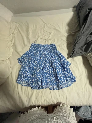Blå volangkjol med vita blommor - Supersöt blå kortkjol med vita blommor och volanger. Kjolen har smock i midjan för skön passform och ett luftigt, flowy tyg som passar perfekt till varma dagar. Perfekt för dig som vill ha en lekfull och trendig look.