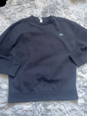 Svart sweatshirt från Lacoste - Klassisk svart sweatshirt från Lacoste med rund halsringning och den ikoniska gröna krokodilloggan broderad på bröstet. Tröjan har långa ärmar och ribbade muddar vid ärmslut och nederkant. Perfekt för en clean och stilren look. REP