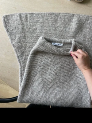 Beige stickad poncho från Adoore - Snygg beige stickad poncho från Adoore med diskret fiskbensmönster. Ponchon har en hög krage och är tillverkad i ett mjukt, värmande material som passar perfekt för lager på lager. Enkel att styla över en tröja eller skjorta för en trendig look.