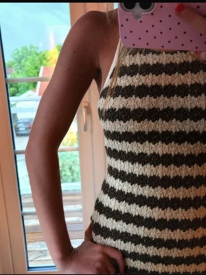 Randig stickad långklänning - Säljer en stickad långklänning med breda beige och mörkbruna ränder. Klänningen har halterneck, öppen rygg med knyt och är ärmlös. Perfekt för sommaren och har en snygg, figurnära passform.