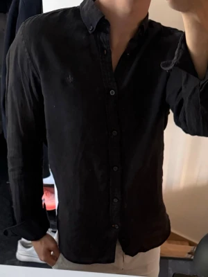 Svart linneskjorta  - Stilren svart linneskjorta med klassisk krage och knappstängning framtill. Skjortan har långa ärmar och en diskret broderad logga på bröstet. Perfekt till jeans eller chinos för en clean look. 100% linne. Lite urtvättad, annars bra skick  