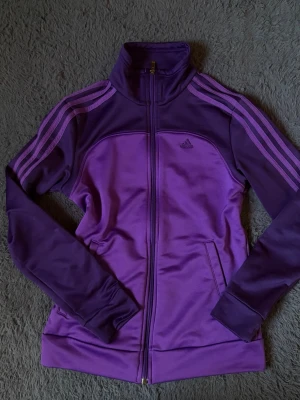 Lila Adidas träningsjacka - Snygg lila träningsjacka från Adidas med klassiska tre ränder på ärmarna och dragkedja framtill. Jackan har hög krage, två fickor och är gjord i ett mjukt, stretchigt material som passar perfekt till träning eller chill. Adidas-logga på bröstet.