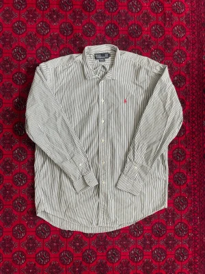 Ralph Lauren Skjorta - Klassisk långärmad skjorta från Polo Ralph Lauren i vit och grön randig bomull. Skjortan har knappar framtill och en liten rosa broderad logga på bröstet. Perfekt för en clean och stilren look.