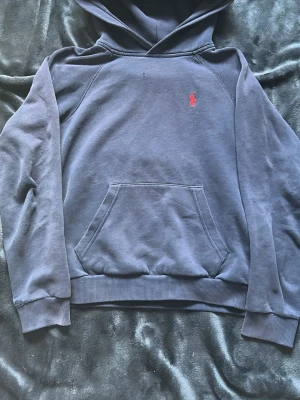 Mörkblå hoodie från Polo Ralph Lauren - Mörkblå hoodie från Polo Ralph Lauren med klassisk röd logga broderad på bröstet. Hoodien har en stor känguruficka framtill, ribbade muddar och mjukt material på insidan. Perfekt för en chill och stilren look.