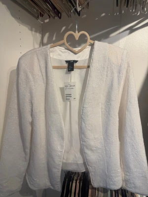 Vit öppen kofta från H&M - Säljer en stilren vit öppen kofta från H&M i storlek L. Koftan har lång ärm, rak passform och ett diskret glittrande material som ger en extra touch. Perfekt att slänga över en topp för en enkel men snygg look.