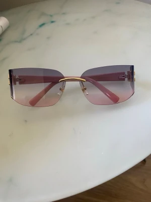 Miu Miu solglasögon rosa/lila - Säljer ett par trendiga solglasögon från Miu Miu med kantlösa, fyrkantiga glas i lila och rosa ton. Skalmarna är rosa med guldfärgade Miu Miu-loggor på sidorna. Snyggt statement-plagg för dig som vill sticka ut.