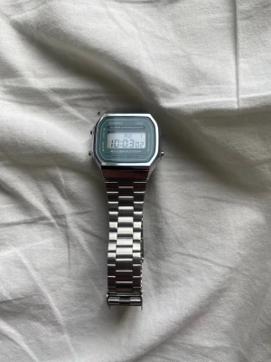 Casio digital klocka i silver - Snygg digital klocka från Casio i klassisk retrostil. Klockan har en rektangulär display med ljusblå bakgrund och silverfärgat armband i rostfritt stål. Funktioner som alarm, datum och belysning. Perfekt accessoar för dig som gillar stilren och tidlös design.