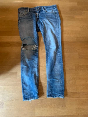 Blå slitna jeans från Polo Ralph Lauren - Säljer ett par blå jeans från Polo Ralph Lauren i 100% bomull. Jeansen har en rak passform, slitningar och hål på vänster knä samt råa kanter vid benslut. Klassisk femficksmodell med knappgylf och tydliga tvättade detaljer. Hör av dig vid minsta fundering!