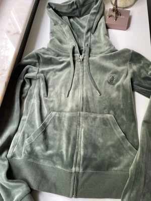 Grön velour hoodie från Juicy Couture - Mjuk och lyxig grön velour hoodie från Juicy Couture med dragkedja, känguruficka och broderad logga på bröstet. Perfekt för en chill och trendig look. Justerbar huva med snören och ribbade muddar för extra komfort.