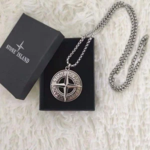 Stone Island halsband med logotyp - Snyggt halsband från Stone Island med en rund, silverfärgad medaljong som har märkets ikoniska kompasslogga och texten 'Stone Island' runt om. Kedjan är grov och metallisk, vilket ger ett coolt och trendigt intryck. Perfekt accessoar för dig som gillar streetwear och vill sticka ut. Skriv innan köp