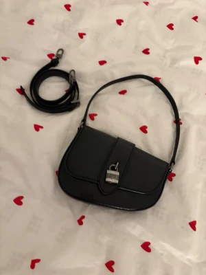Svart handväska från Guess - Snygg svart handväska från Guess i glansigt konstläder med subtilt ormmönster. Väskan har ett dekorativt hänglås framtill med Guess-logga och gnistrande stenar. Justerbar axelrem medföljer så du kan bära den både på axeln och crossbody.