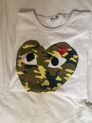 Vit Comme des Garçons PLAY t-shirt - Vit t-shirt från Comme des Garçons PLAY med stort hjärta i camouflage-mönster och ett litet rött hjärta med ögon på bröstet. Klassisk rund hals och korta ärmar. Perfekt statement-plagg för dig som gillar streetwear.
