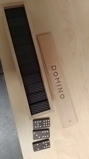 Dominoset i trä med bruna brickor - Säljer ett klassiskt dominoset i en ljus trälåda med texten DOMINO på locket. Brickorna är mörkbruna med vita prickar och har en räfflad yta. Perfekt för spelkvällar med vänner eller familj.