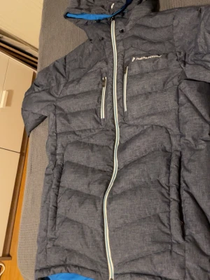 Grå pufferjacka Peak Performance S - Snygg grå pufferjacka från Peak Performance med blått innerfoder och vita detaljer. Jackan har huva, två bröstfickor med dragkedja och hel dragkedja framtill. Perfekt för kalla dagar och har en modern, quiltad look.