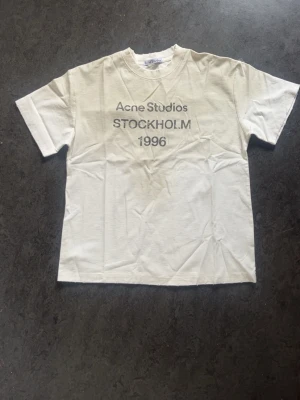 Vit Acne Studios t-shirt Stockholm 1996 - Vit t-shirt från Acne Studios med tryck 'STOCKHOLM 1996' på bröstet. Klassisk rak passform och rund halsringning. Perfekt basic-plagg med stilren design och diskret logga. Tillverkad i mjuk bomullsblanding. Kvitto finns