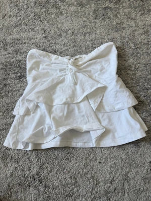 Vit bandeau topp från Gina Tricot - Säljer en vit bandeau topp från Gina Tricot med volangdetaljer och knytning framtill. Toppen är axelbandslös och har en snygg lager-på-lager look som ger extra volym. Perfekt för sommarens alla outfits!