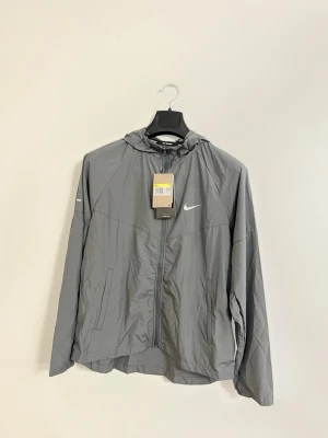 Nike Miller Track Suit Grå – Storlek S - Säljer en helt ny Nike Miller Track Suit i grå färg, storlek S. Setet består av en lätt jacka med huva och matchande byxor. Perfekt för träning, löpning eller en sportig vardagsstil. 